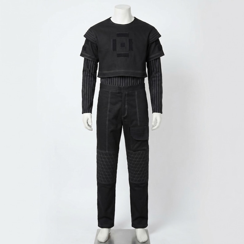 Op de afbeelding: Zwart ensemble op een mannequin. Het bevat een crop top met korte mouwen en een geometrisch ontwerp, een gestreept shirt met lange mouwen en een zwarte broek met gewatteerde kniepanelen en zakdetails.