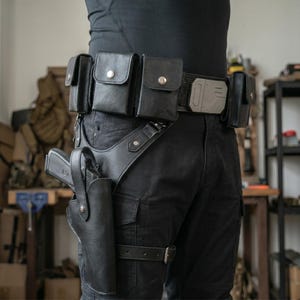 Op de afbeelding: Zwarte leren utility riem met meerdere zakjes en een holster. De riem heeft een zilverkleurige gesp en een beenriem. De holster bevat een zwart pistool. De riem wordt gedragen met een zwarte broek.