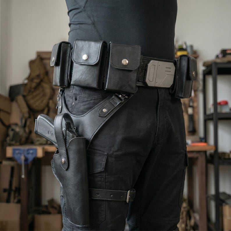 Op de afbeelding: Zwarte leren utility riem met meerdere zakjes en een holster. De riem heeft een zilverkleurige gesp en een beenriem. De holster bevat een zwart pistool. De riem wordt gedragen met een zwarte broek.