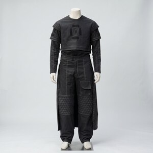 Op de afbeelding: Een zwart ensemble op een mannequin. De outfit bestaat uit een top met korte mouwen en een geometrisch ontwerp, een shirt met lange mouwen eronder en een broek met wijde pijpen, gewatteerde kniedetails en een zijrok. Witte stiksels accentueren de outfit.