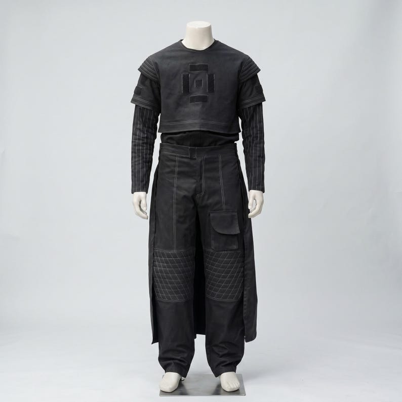 Op de afbeelding: Een zwart ensemble op een mannequin. De outfit bestaat uit een top met korte mouwen en een geometrisch ontwerp, een shirt met lange mouwen eronder en een broek met wijde pijpen, gewatteerde kniedetails en een zijrok. Witte stiksels accentueren de outfit.