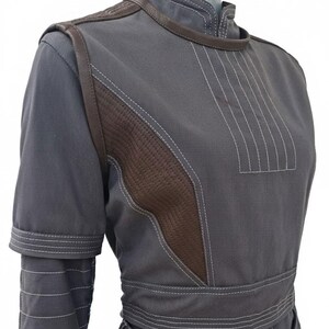 May include: A gray jumpsuit with brown leather accents and white stitching. The jumpsuit features a high collar, padded shoulders, and a fitted waist. The brown leather is quilted on the chest and shoulder areas. The sleeves have a layered design.