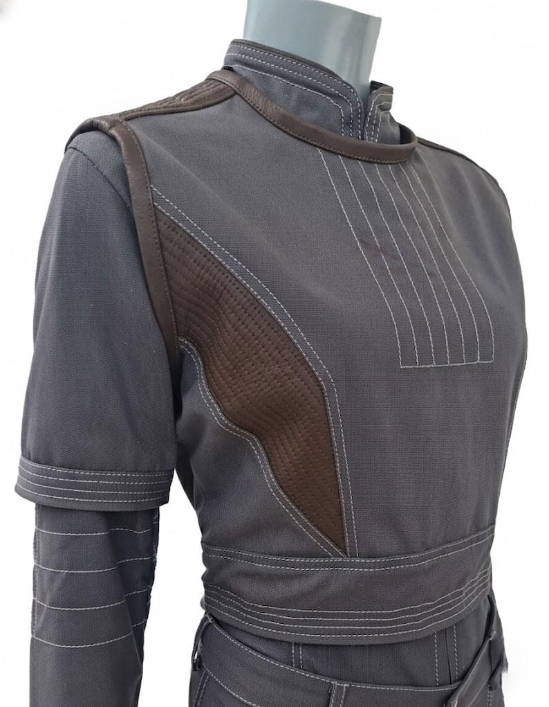 May include: A gray jumpsuit with brown leather accents and white stitching. The jumpsuit features a high collar, padded shoulders, and a fitted waist. The brown leather is quilted on the chest and shoulder areas. The sleeves have a layered design.