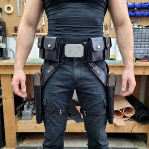 Könnte beinhalten: Schwarzer Leder-Werkzeuggürtel mit mehreren Taschen, einer silbernen Schnalle und Beingurten. Der Gürtel hat zwei Holster und ist für das Tragen von Werkzeugen oder Zubehör konzipiert. Der Gürtel wird über einer dunkelblauen Jeans getragen.