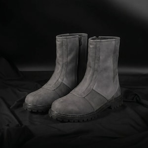 May include: A pair of gray ankle boots with a rugged, textured appearance. The boots feature a zipper closure and a two-tone design, with a darker gray panel at the base. The soles are black and have a treaded design.