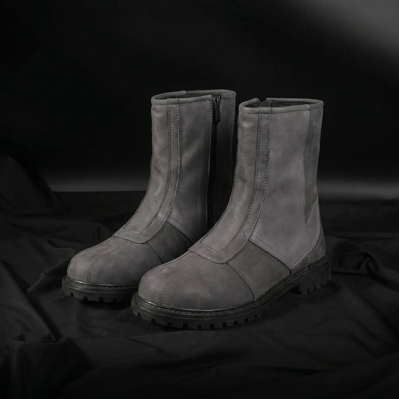 May include: A pair of gray ankle boots with a rugged, textured appearance. The boots feature a zipper closure and a two-tone design, with a darker gray panel at the base. The soles are black and have a treaded design.