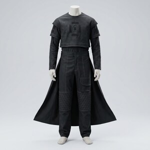 Op de afbeelding: Een zwarte outfit op een mannequin, bestaande uit een top met korte mouwen met een geometrisch ontwerp, een gestreept ondershirt met lange mouwen, een broek met hoge taille en een lange jas. De broek heeft gewatteerde knielappen. De outfit is gemaakt van mat materiaal.