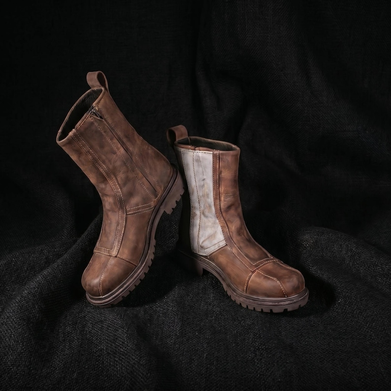 K&ouml;nnte beinhalten: Zwei braune Lederstiefel mit Rei&szlig;verschluss und Zuglasche. Ein Stiefel hat eine wei&szlig;e Seite. Die Stiefel haben eine robuste Sohle und sind vor einem dunklen Hintergrund platziert.