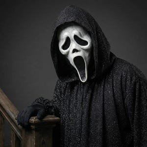 May include: A person wearing a black hooded robe and a white Scream mask with a wide open mouth. The person is wearing black gloves and is standing near a wooden banister. The robe has a glittery texture.