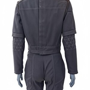 May include: A dark gray jumpsuit with white stitching details. The jacket has a cropped design with geometric patterns on the sleeves. The pants are fitted, and the outfit has a structured, tailored appearance.