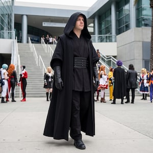May include: A full-length black hooded cloak worn over a black tunic and matching trousers. The outfit includes a wide black belt with horizontal stitching, black leather gloves, and black dress shoes, creating a monochromatic dark ensemble.