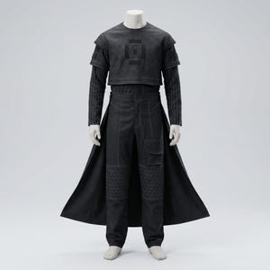 Op de afbeelding: Een zwarte outfit op een mannequin, bestaande uit een top met korte mouwen met een geometrisch ontwerp, een gestreept ondershirt met lange mouwen, een broek met hoge taille en een lange jas. De broek heeft gewatteerde knielappen. De outfit is gemaakt van mat materiaal.