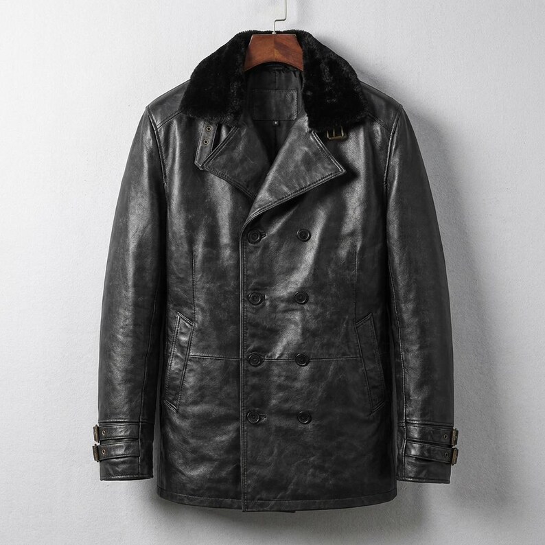K&ouml;nnte beinhalten: Ein schwarzer Ledercoat mit doppelreihigem Design und einem Kunstfellkragen. Der Mantel hat zwei Knopfreihen, zwei Vordertaschen und verstellbare Riemen an den &Auml;rmeln. Der Mantel h&auml;ngt an einem Holzb&uuml;gel.