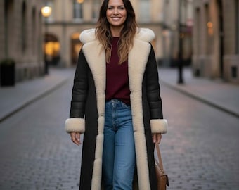 May include: A long black coat with cream-colored shearling trim and lining. The coat features a hood and is worn over a burgundy sweater and blue jeans. A brown purse is carried over the shoulder. The person is walking down a cobblestone street.
