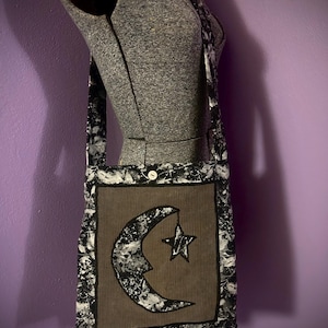 May include: A black and white fabric tote bag with a brown crescent moon and star design. The bag has a long strap and a button closure.
