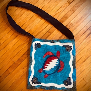May include: A blue and white patchwork tote bag with a red and white turtle design. The turtle has a Grateful Dead Steal Your Face logo on its shell.