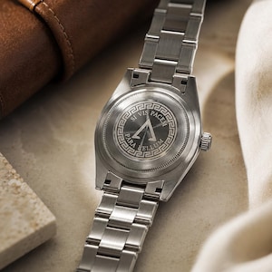 May include: A silver-toned stainless steel wristwatch with a metal link bracelet. The circular case back features an engraved Greek key border surrounding a central triangle symbol and the Latin text SI VIS PACEM PARA BELLUM.