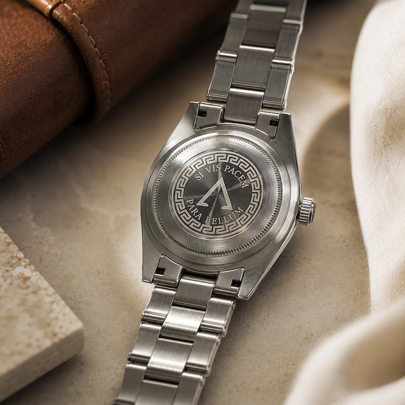 May include: A silver-toned stainless steel wristwatch with a metal link bracelet. The circular case back features an engraved Greek key border surrounding a central triangle symbol and the Latin text SI VIS PACEM PARA BELLUM.