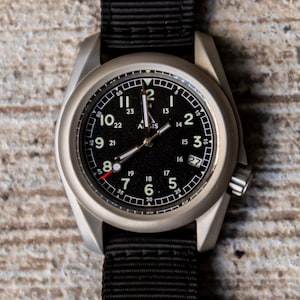 以下が含まれることがあります： 黒い文字盤と黒いナイロンバンドが付いた、シルバーの腕時計。 文字盤には白い数字とマーカーが表示され、3時の位置に日付表示があります。 時計はクラシックで実用的なデザインです。