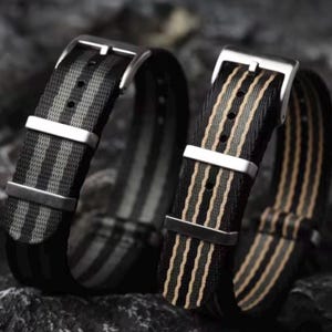 May include: Two nylon watch straps featuring a striped pattern. The left strap has black and gray stripes, while the right strap has black and tan stripes. Both straps include a silver-toned metal buckle and two matching silver-toned metal keepers.