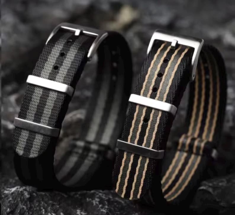 May include: Two nylon watch straps featuring a striped pattern. The left strap has black and gray stripes, while the right strap has black and tan stripes. Both straps include a silver-toned metal buckle and two matching silver-toned metal keepers.