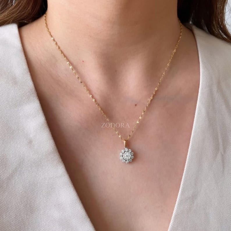 Puede incluir: Un collar de cadena dorada con un colgante redondo que presenta una piedra central transparente rodeada por un halo de piedras m&aacute;s peque&ntilde;as. El colgante cuelga de una delicada cadena de eslabones dorados. La palabra ZODORA es visible arriba.