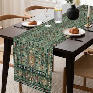 Peut inclure: Un chemin de table vert à motifs avec un design détaillé, placé sur une table en bois sombre. La table est dressée avec des assiettes, des couverts, un verre et une carafe d'eau. Un vase noir avec des fleurs blanches et une bougie allumée sont également présents.