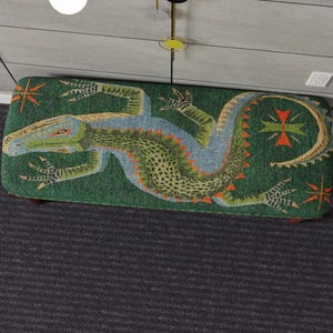 May include: A rectangular upholstered bench with a green background and a large lizard design. The lizard is primarily green, blue, and orange. The bench has dark wooden legs and decorative starburst patterns.