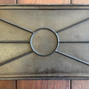 Op de afbeelding: Rechthoekige gietijzeren utility grill met een draadrek inzetstuk. De grill heeft handvatten aan elk uiteinde en de woorden "Wagner Ware Sidney -0- UTILITY GRILL" zijn in reliëf. De grill is donkergrijs en ligt op een houten oppervlak.