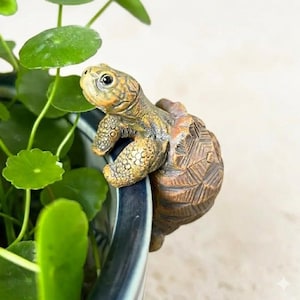 Pode incluir: Uma detalhada estatueta de tartaruga marrom e bronzeada pousada na borda de um vaso de cerâmica azul e branco. A tartaruga tem uma carapaça texturizada e detalhes precisos. Plantas verdes e frondosas ao fundo.