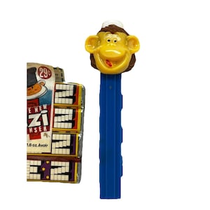 May include: A vintage PEZ dispenser featuring a smiling monkey head with a white hat, atop a blue dispenser. The packaging shows a cartoon monkey and the text "PEZ" and "Zipper". The packaging also has the text "1.8 oz Avoir".