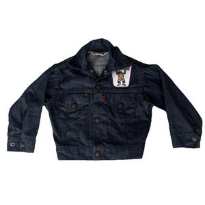 Puede incluir: Chaqueta vaquera azul oscuro con un parche con un personaje de dibujos animados. La chaqueta tiene cierre de botones, dos bolsillos en el pecho y puños abotonados. La etiqueta de la marca Levi's es visible dentro del cuello.