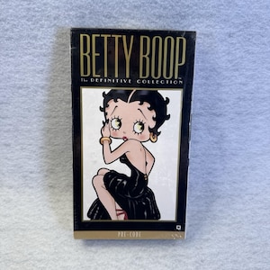 Könnte beinhalten: Eine rechteckige DVD-Hülle von Betty Boop The Definitive Collection mit schwarzem Hintergrund und goldenem Text. Die zentrale Illustration zeigt die Zeichentrickfigur Betty Boop mit schwarzen Haaren in einem schwarzen Kleid vor weißem Hintergrund.