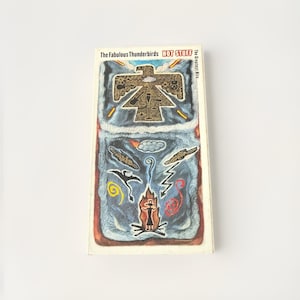 Puede incluir: Una portada de álbum vintage de "The Fabulous Thunderbirds: Hot Stuff". La obra de arte presenta un diseño estilizado de Thunderbird nativo americano en oro sobre un fondo azul y gris, con ilustraciones coloridas adicionales.