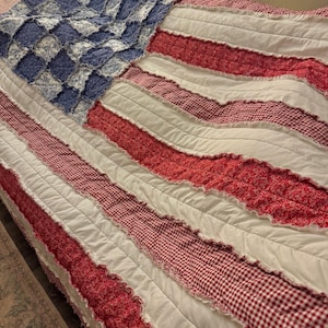 May include: A handmade quilt in the shape of the American flag. The quilt features a blue square with white floral patterns, red and white gingham stripes, and white stripes. The quilt is made of soft fabric and is likely for a bed.