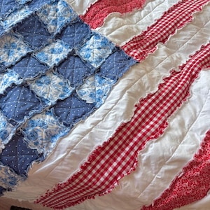 Könnte beinhalten: Eine handgefertigte Steppdecke mit patriotischem Design. Die Steppdecke zeigt ein blaues Quadrat mit Blumen- und Punktmustern, das die Sterne darstellt. Rote und weiße Streifen, einige mit Vichy-Muster, vervollständigen das Thema der amerikanischen Flagge.