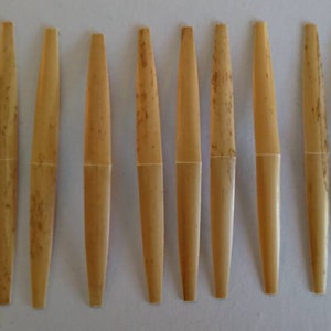 May include: Eight natural brown reed oboe reeds, arranged in a row on a white surface.