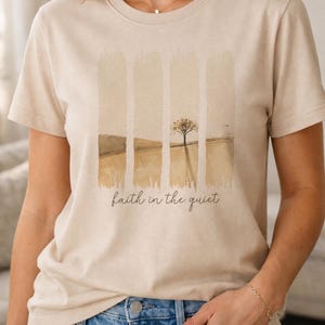 May include: A beige short-sleeved t-shirt featuring a graphic print of a landscape with a single tree divided into four vertical panels. Below the graphic, the text "faith in the quiet" is printed in a dark brown, cursive script font.