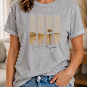 Op de afbeelding: Een lichtgrijs gem&ecirc;leerd t-shirt met korte mouwen en een grafische print van een landschap verdeeld in vier verticale panelen. Het ontwerp toont een goudbruin veld en een enkele boom, met de tekst "faith in the quiet" eronder geschreven.
