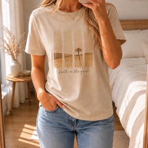 Op de afbeelding: Een beige t-shirt met korte mouwen en een grafische print van een landschap verdeeld in vier verticale panelen. Het ontwerp toont een eenzame boom in een veld met de tekst "faith in the quiet" in een cursief lettertype eronder.
