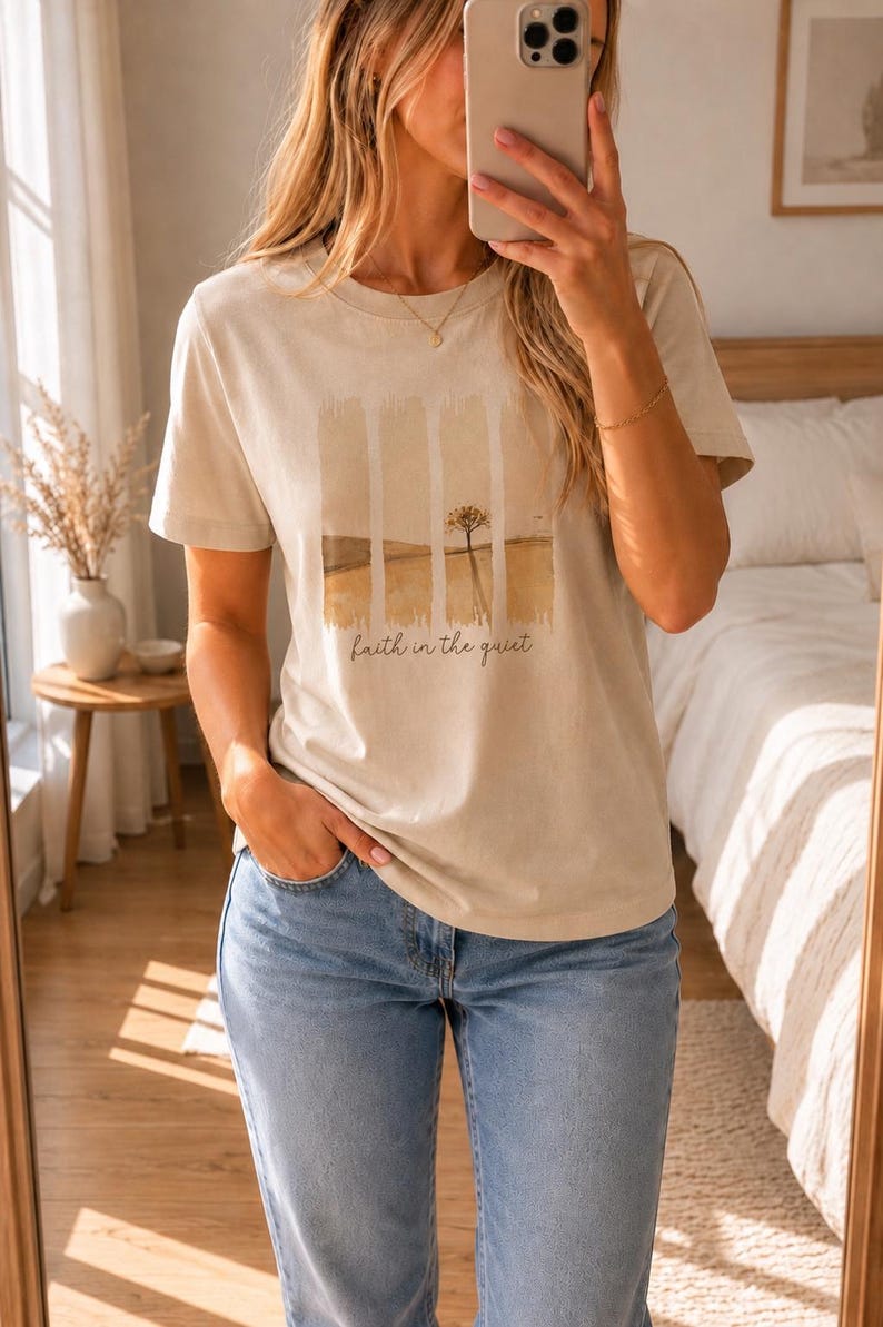 Op de afbeelding: Een beige t-shirt met korte mouwen en een grafische print van een landschap verdeeld in vier verticale panelen. Het ontwerp toont een eenzame boom in een veld met de tekst "faith in the quiet" in een cursief lettertype eronder.