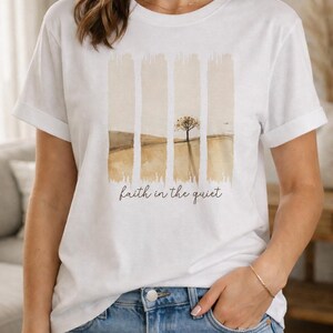 Op de afbeelding: Een wit t-shirt met korte mouwen en een afbeelding van vier verticale penseelstreken die een landschap met een enkele boom in gedempte bruine en beige tinten weergeven. De tekst "faith in the quiet" staat in een donkerbruin cursief lettertype.