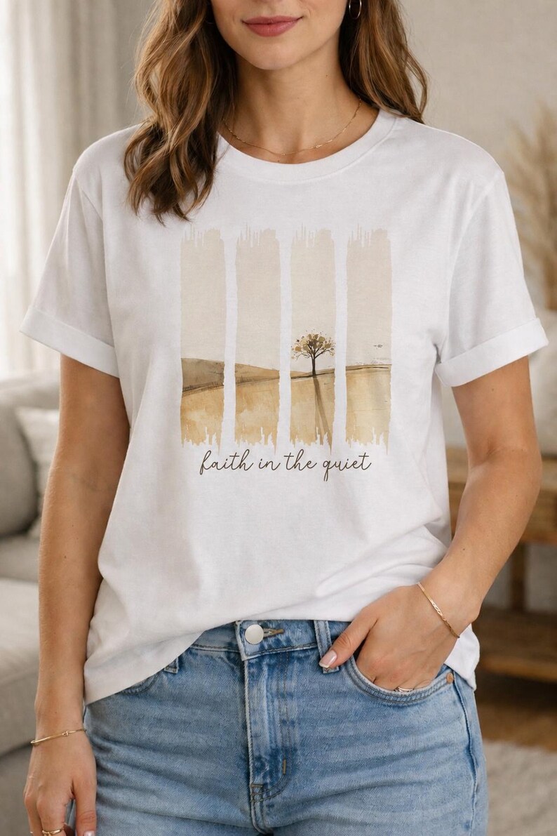 Op de afbeelding: Een wit t-shirt met korte mouwen en een afbeelding van vier verticale penseelstreken die een landschap met een enkele boom in gedempte bruine en beige tinten weergeven. De tekst "faith in the quiet" staat in een donkerbruin cursief lettertype.