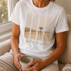 Op de afbeelding: Een wit T-shirt met korte mouwen en een grafische print van een landschap met een enkele boom en heuvels in beige en bruine tinten. Het ontwerp is verdeeld door verticale penseelstreken en bevat de tekst faith in the quiet eronder.