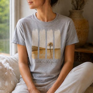 Op de afbeelding: Een grijs gem&ecirc;leerd t-shirt met korte mouwen en een grafische print van een eenzame boom in een landschap met verticale penseelstreekaccenten. De tekst "faith in the quiet" staat in cursief schrift onder de landschapsillustratie gedrukt.