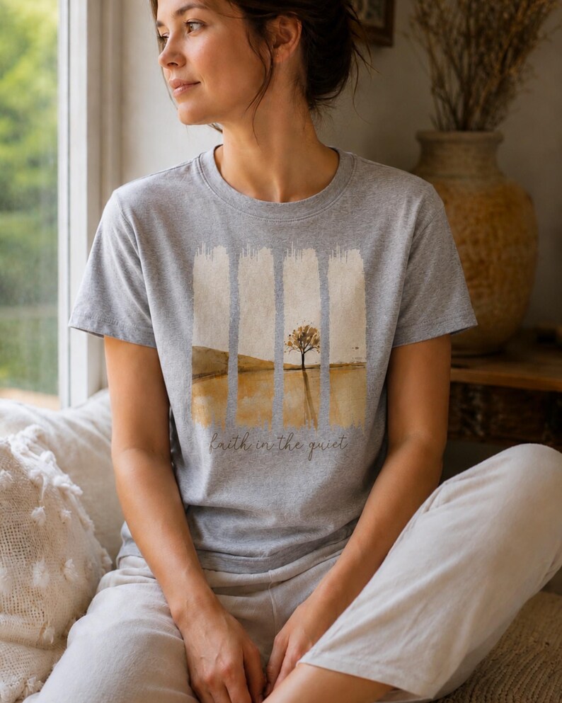 Op de afbeelding: Een grijs gem&ecirc;leerd t-shirt met korte mouwen en een grafische print van een eenzame boom in een landschap met verticale penseelstreekaccenten. De tekst "faith in the quiet" staat in cursief schrift onder de landschapsillustratie gedrukt.