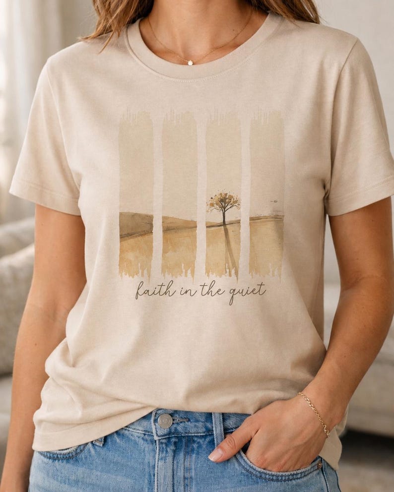 May include: A beige short-sleeved t-shirt featuring a graphic print of a landscape with a single tree divided into four vertical panels. Below the graphic, the text "faith in the quiet" is printed in a dark brown, cursive script font.