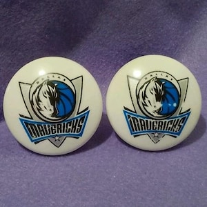 May include: Two white round buttons featuring the Dallas Mavericks logo. The logo includes a stylized horse head, a basketball, and the word "MAVERICKS" in blue and white. The buttons are set against a purple background.