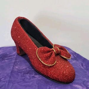 May include: A single red glitter-covered shoe with a bow and a small heel. The bow is outlined with gold trim. The shoe is on a purple surface.