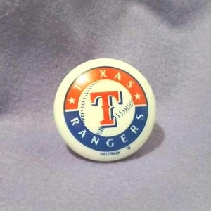 Peut inclure: Un bouton rond blanc avec le logo de l'équipe de baseball des Texas Rangers. Le logo comprend un "T" rouge avec une balle de baseball, entouré des mots "TEXAS RANGERS" en rouge et bleu, avec des étoiles et un bord blanc.